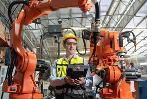 A worker operates a control panel between two industrial robotic arms in a factory setting